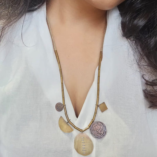 A person wearing a white shirt models a geometric necklace with oxidized brass patterns. The necklace features brass, copper, and jute thread.
