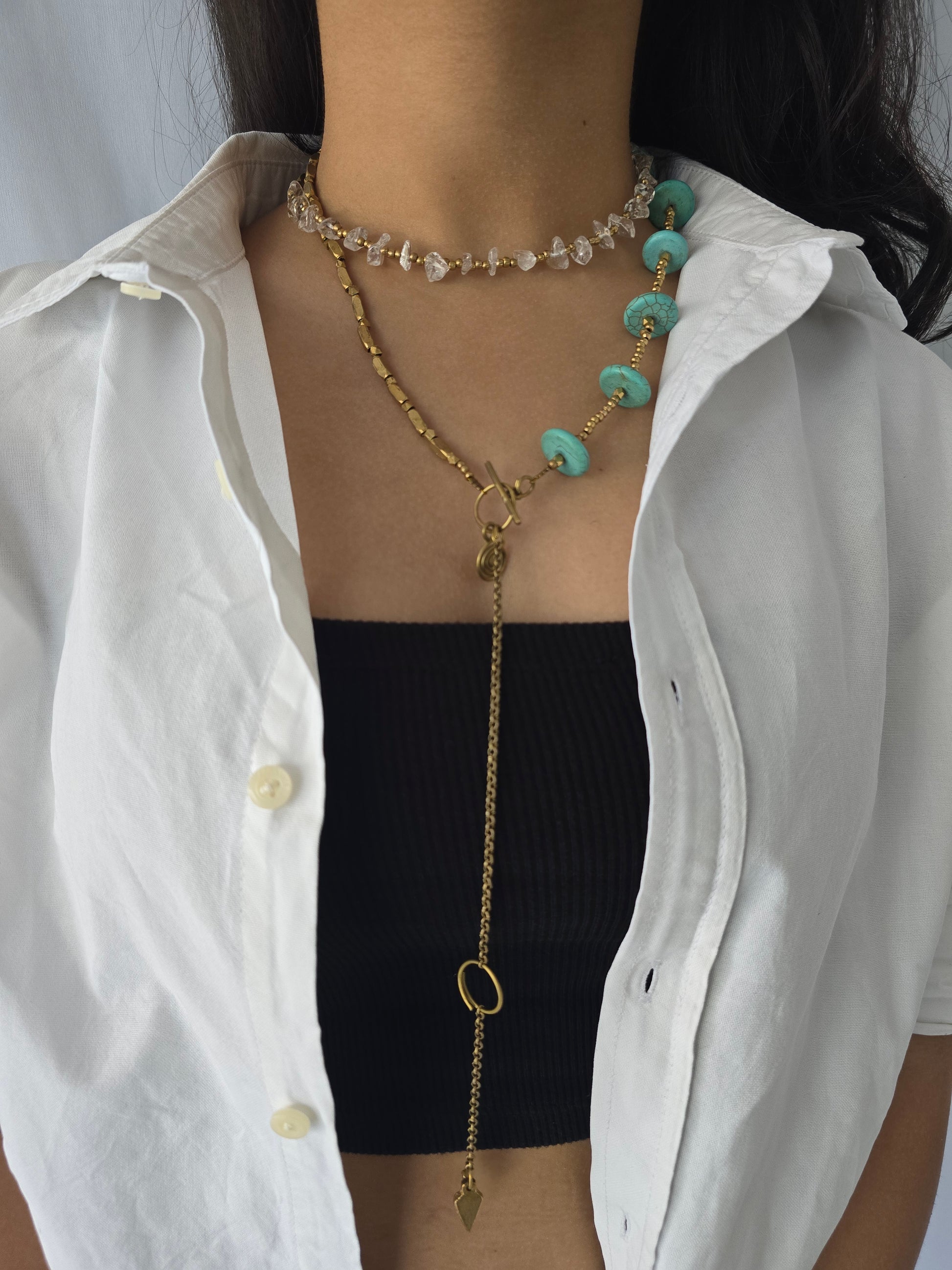 A person wearing a white shirt and black top, adorned with a gold-toned necklace featuring brass beads, turquoise howlite, and clear quartz in a wrap-around style.