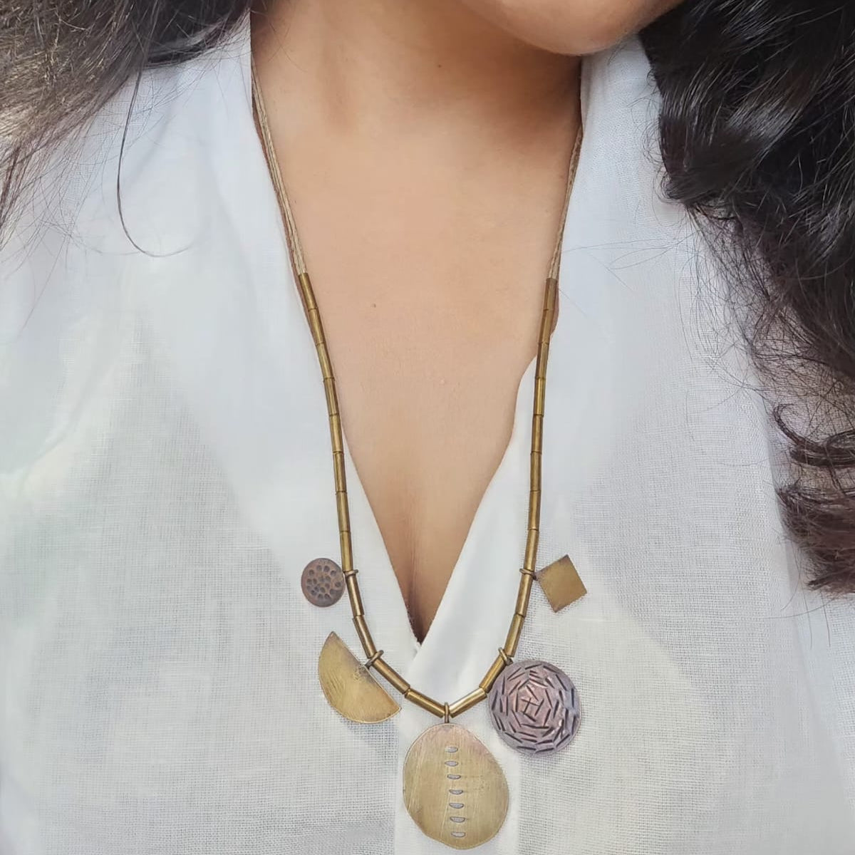 A person wearing a white shirt models a geometric necklace with oxidized brass patterns. The necklace features brass, copper, and jute thread.