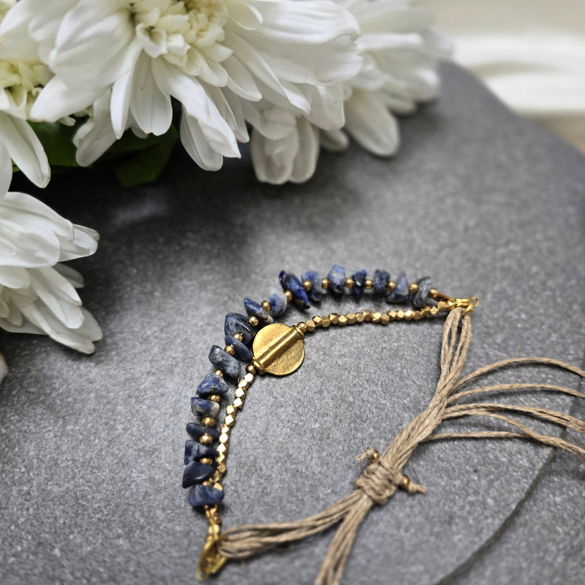 Lumba bracelet with Lapis Lazuli beads and gold accents on a gray surface with white flowers.