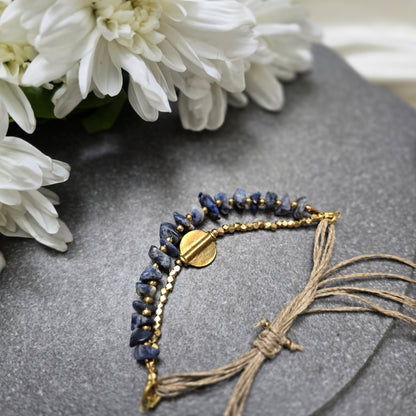 Lumba bracelet with Lapis Lazuli beads and gold accents on a gray surface with white flowers.